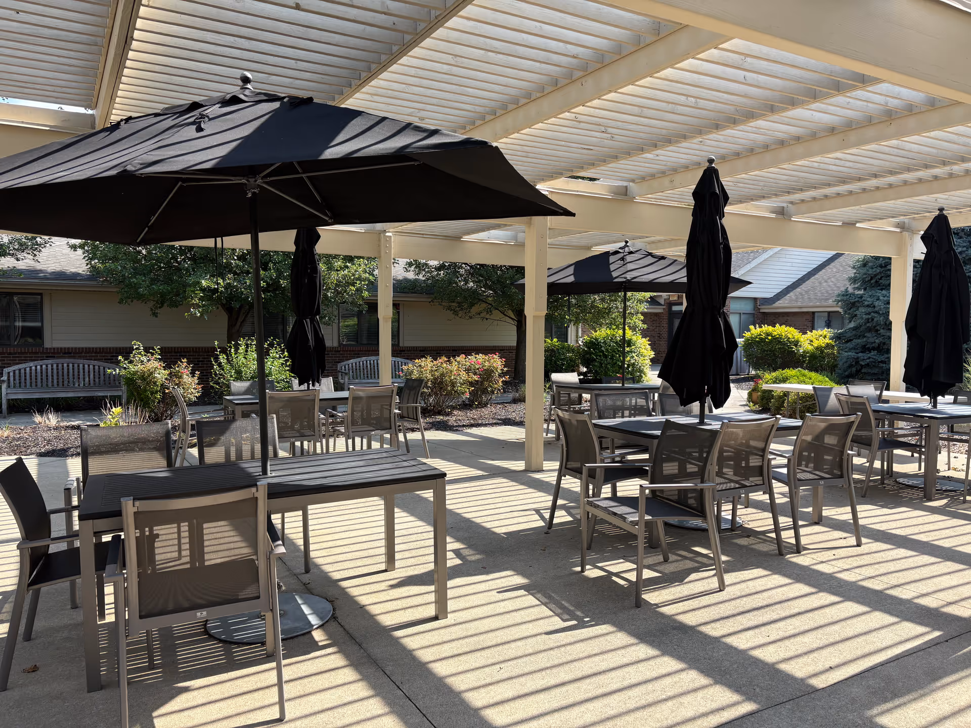 Shaded outdoor patio with tables, chairs, umbrellas, and benches under a pergola next to landscaped grounds and a building.