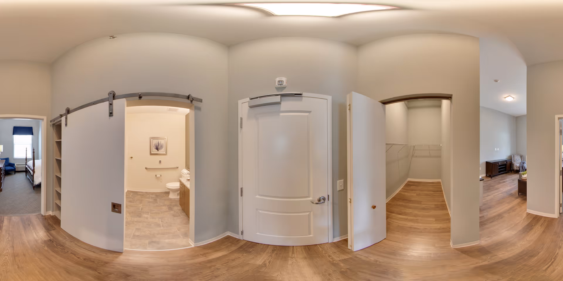 Interior view of a hallway in an assisted living facility showing multiple doorways. On the left, there is a bedroom with a bed and window. In the center, a bathroom with a toilet, grab bars, and a framed picture on the wall is visible through a sliding barn door. To the right, there is an open door leading to a walk-in closet with shelving. The hallway has wood flooring and neutral-colored walls with soft lighting.