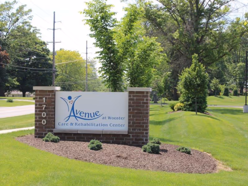 Brick entrance sign for the Avenue at Wooster Care & Rehabilitation Center on a landscaped lawn with trees and a road in the background.