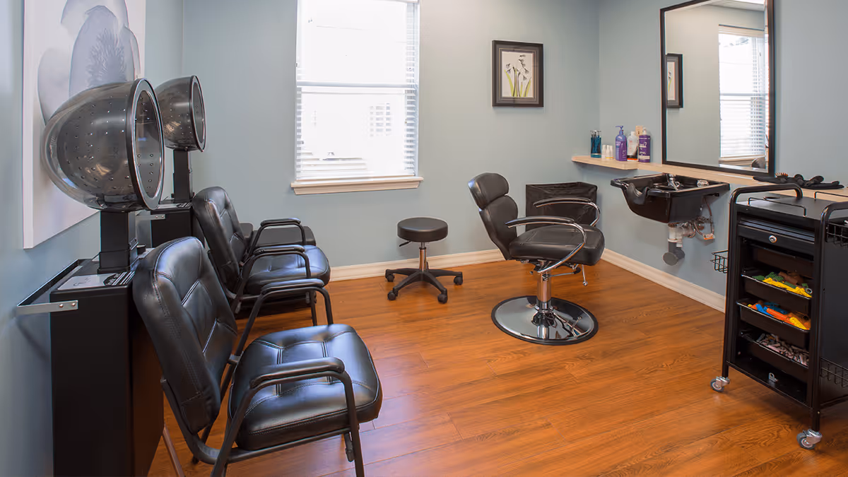 Interior of a hair salon area with two black hair dryer chairs on the left, a black salon chair in front of a mirror and sink on the right, a small black rolling stool in the center, and a black rolling cart with hair products and tools. The room has light blue walls, wooden flooring, a window with blinds, and a framed picture on the wall.