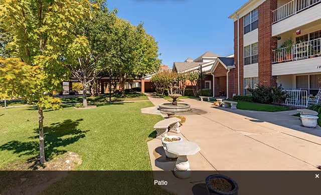 Outdoor patio area at Broadway Cityview featuring a paved walkway, stone benches, a central water fountain, green grass, and trees with a multi-story brick building in the background under a clear blue sky.