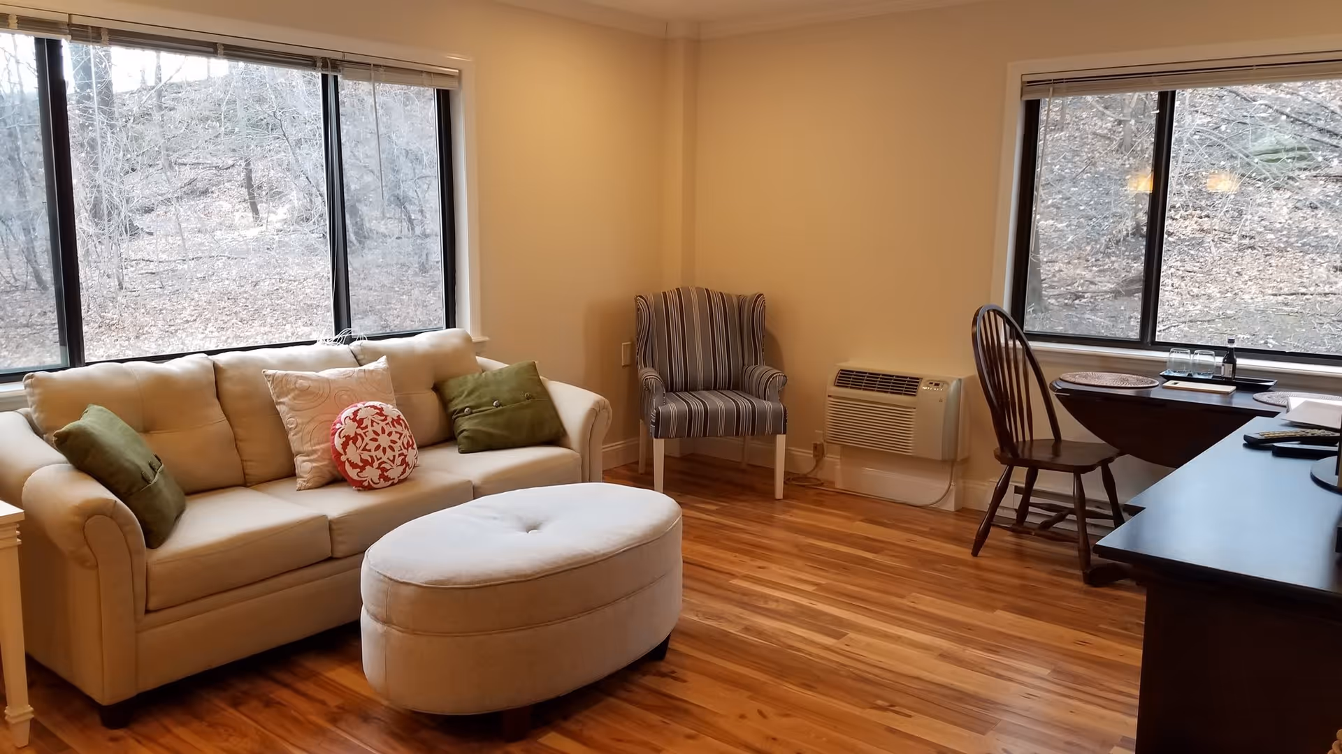 A cozy living room with a beige sofa adorned with green and patterned pillows, a matching oval ottoman, a striped armchair, and a wooden chair next to a small table. The room has large windows showing a view of leafless trees outside and a wooden floor.
