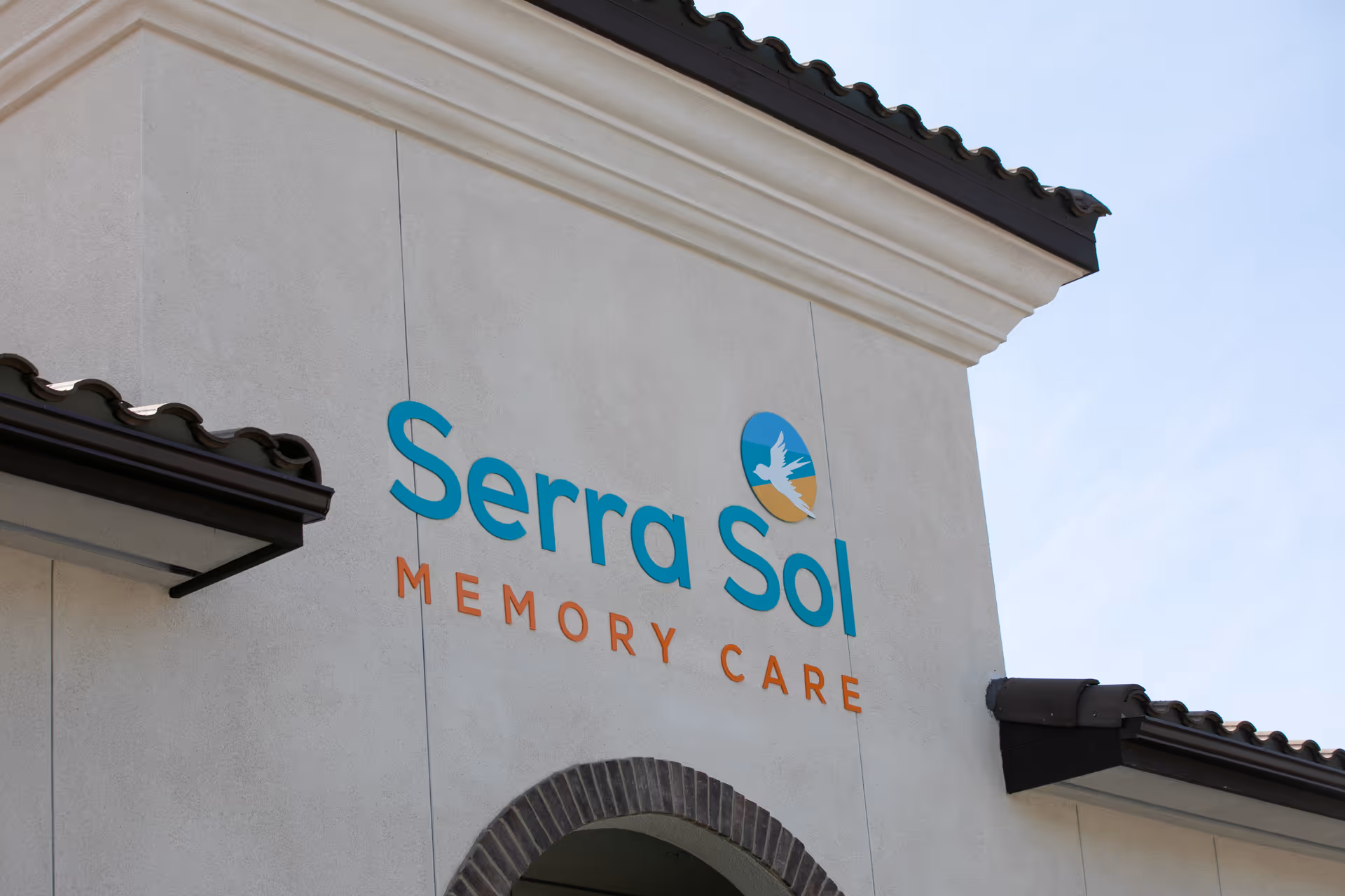 Exterior view of a building wall with the sign 'Serra Sol Memory Care' mounted on it, featuring a logo with a bird flying over a sun and hills.