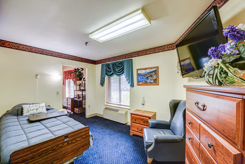 A cozy bedroom in a senior living facility featuring a single bed with a gray comforter and a pillow that says 'live simply'. The room has a blue carpet, a wooden dresser with a flat-screen TV mounted above it, a comfortable armchair, a small wooden nightstand with a lamp, and a window with teal curtains. There is a decorative painting on the wall and an air conditioning unit below the window.