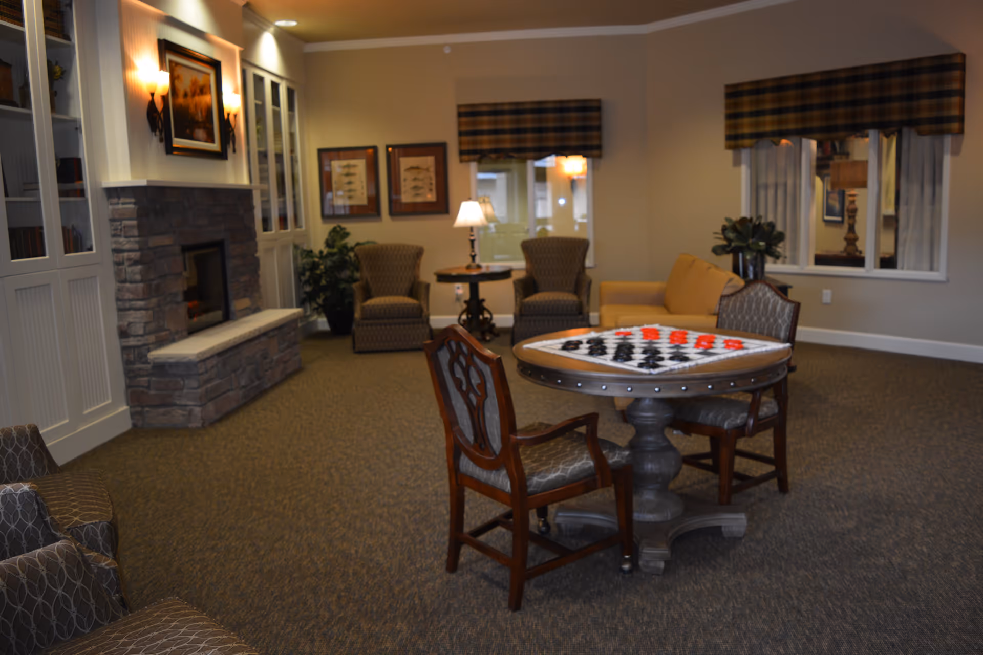 A cozy living room area with a stone fireplace on the left, two armchairs and a small table with a lamp near the back wall, a yellow couch, and a round table with a checkerboard game set up. The room has carpeted flooring, framed artwork on the walls, and windows with plaid valances.