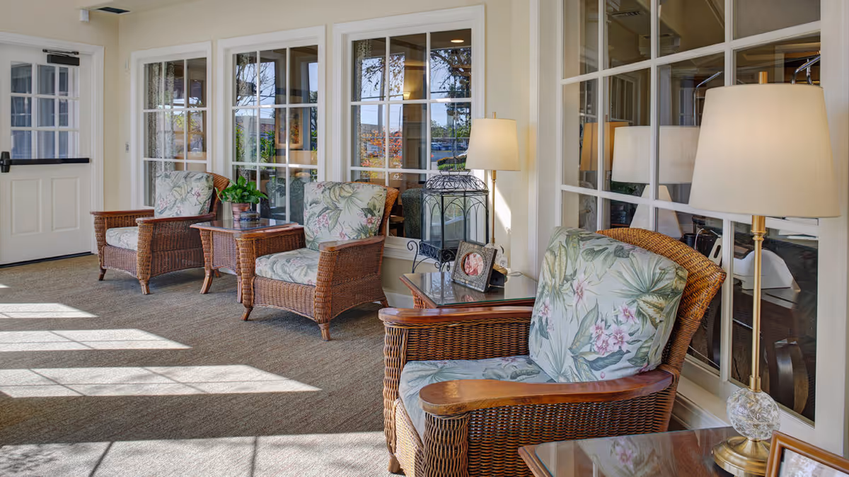 A sunlit seating area with three wicker armchairs featuring floral cushions, each paired with a small wooden side table. The space has large windows with white frames, allowing natural light to fill the room. A floor lamp with a white shade is positioned next to one of the chairs, and decorative items including a framed photo and a plant are on the tables.