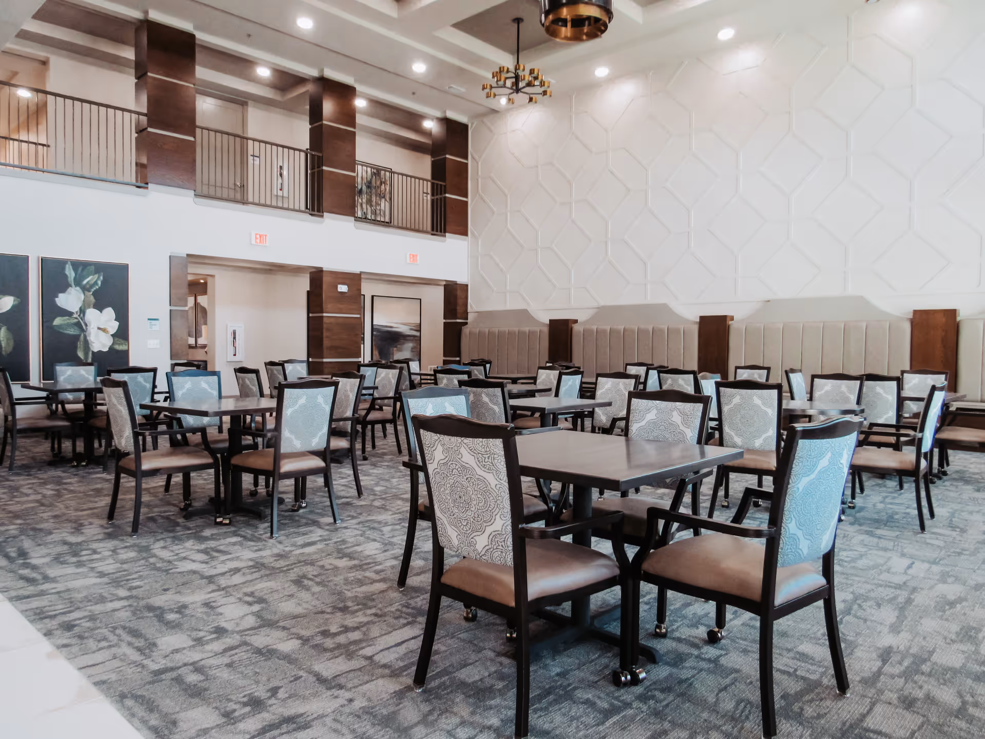 Spacious dining room with multiple square tables and upholstered chairs arranged across a carpeted floor under a high decorative wall.