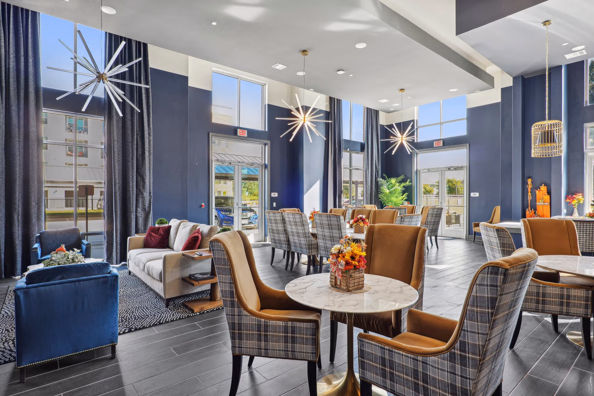 A bright and modern common area with high ceilings and large windows. The room features multiple seating arrangements including plaid and tan upholstered chairs around small round tables with floral centerpieces, a beige sofa with red pillows, and blue armchairs on a patterned rug. Contemporary starburst pendant lights hang from the ceiling, and there are large glass doors leading outside.