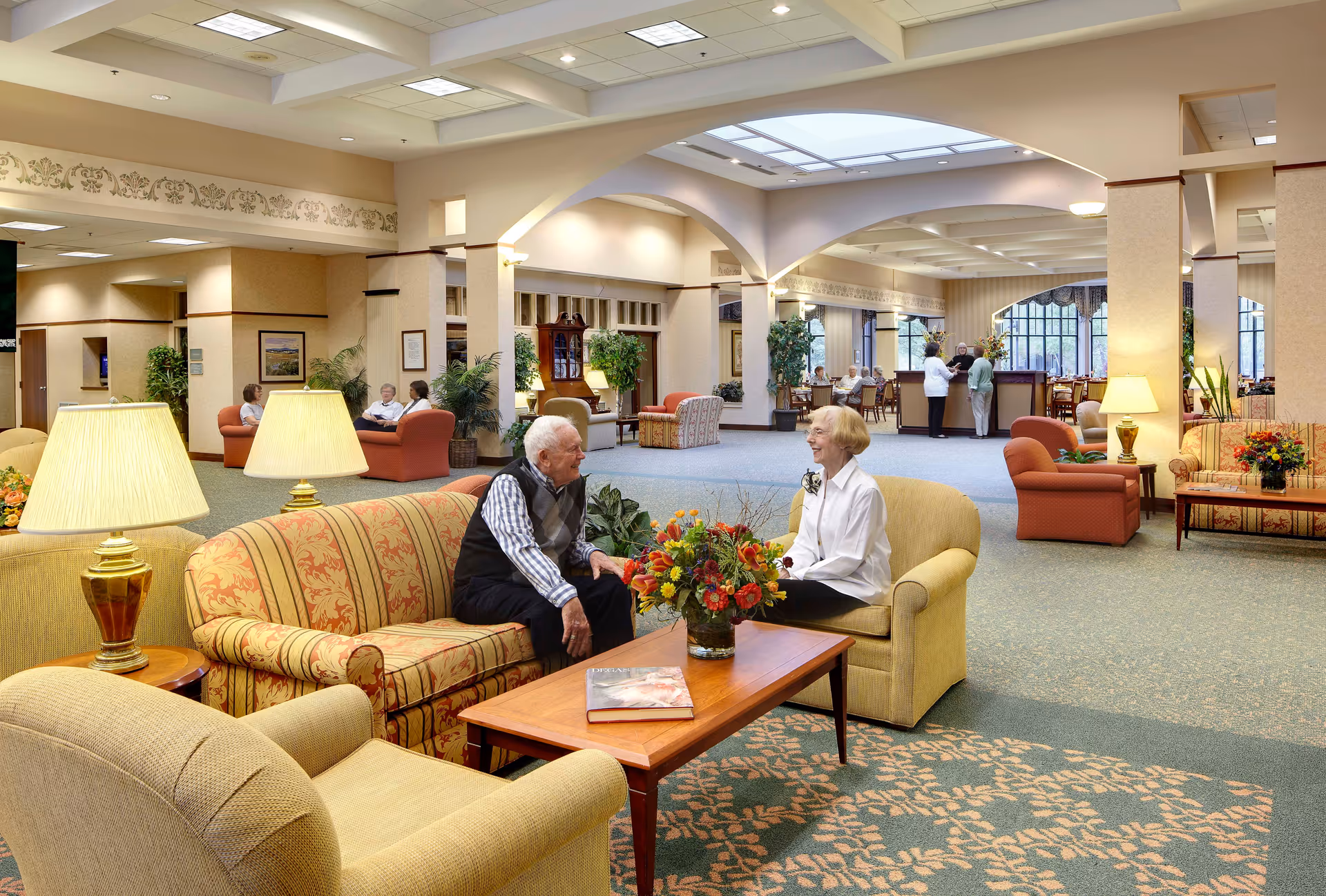 Spacious senior living facility lounge with sofas, armchairs, lamps, and residents conversing around a coffee table.
