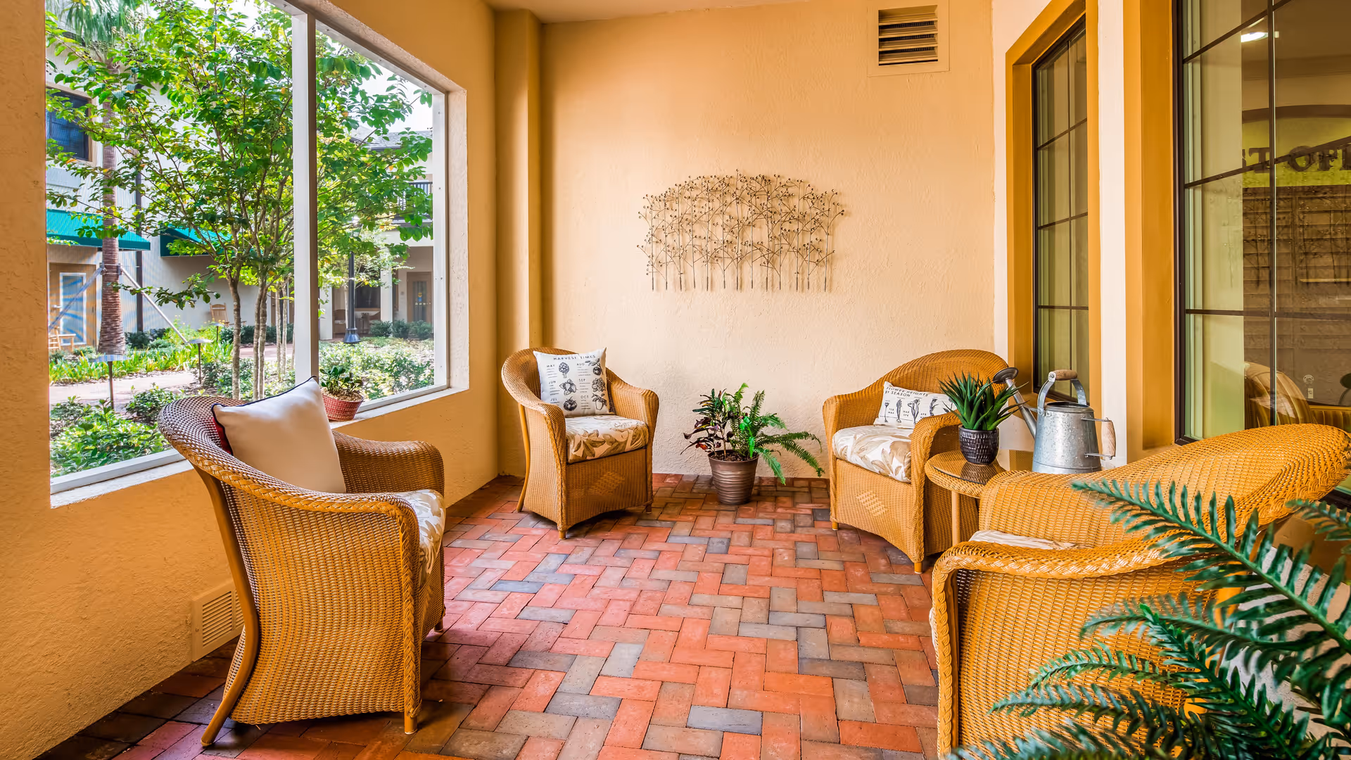 A cozy indoor seating area with four wicker chairs arranged around a small table. The floor is covered with red and gray brick tiles in a herringbone pattern. There are potted plants on the table and on the floor, and a decorative metal wall art piece hangs on the beige wall. Large windows provide a view of green trees and plants outside.