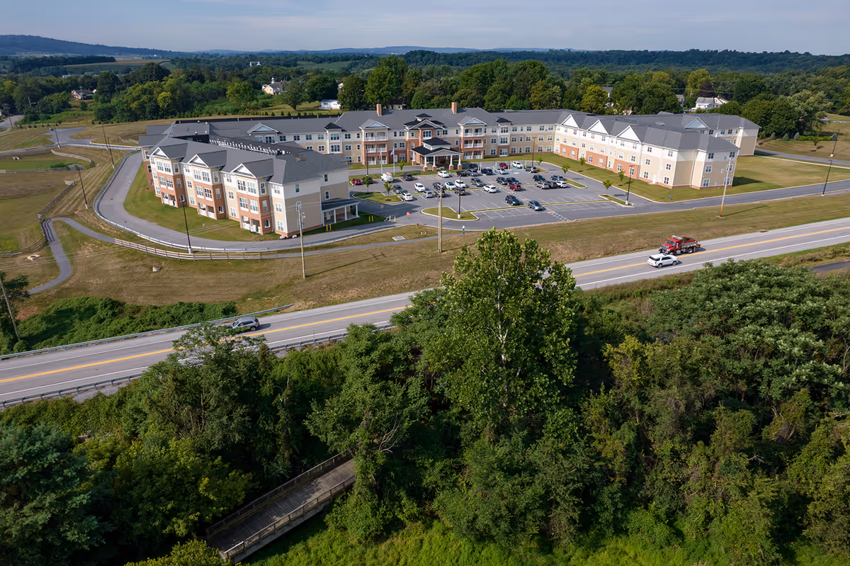 Aerial view of Harmony at Hershey senior living facility showing a large, multi-story building with a parking lot in front, surrounded by green trees and open land, with a road running parallel to the building.