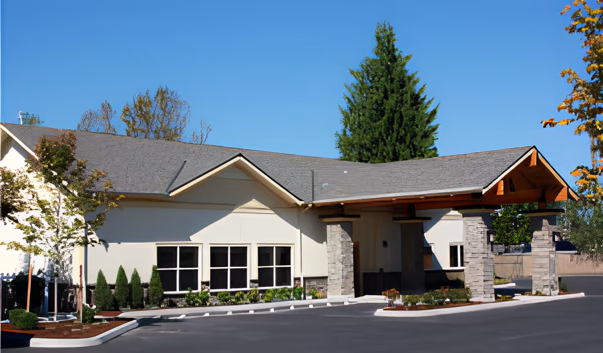 Exterior view of a single-story assisted living facility building with a covered entrance supported by stone pillars, surrounded by landscaped greenery and trees under a clear blue sky.