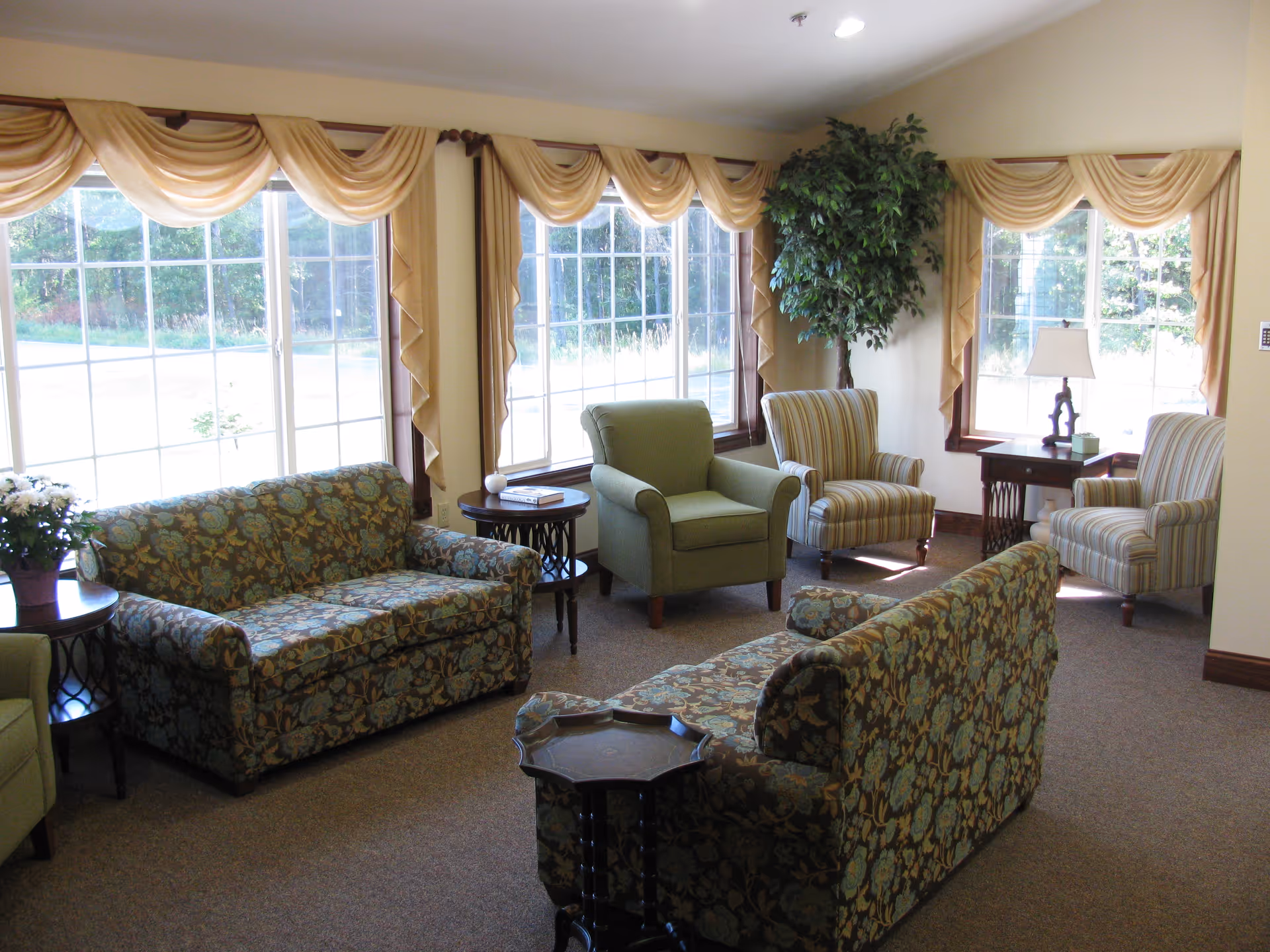 A bright and cozy living room with large windows covered by beige drapes. The room contains two floral patterned sofas, two striped armchairs, one solid green armchair, several small wooden side tables, a table lamp, a potted plant, and a vase of flowers. The carpet is brown and the walls are light-colored.