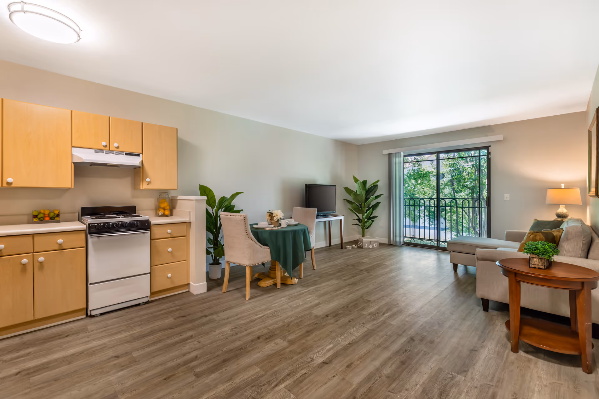 A bright and spacious studio apartment with a small kitchen area featuring light wood cabinets and a white stove. There is a small round dining table with two upholstered chairs covered with a green tablecloth. The living area has a beige sectional sofa with green and brown pillows, a wooden side table with a small plant, and a TV on a stand. Large sliding glass doors lead to a balcony with a black railing and a view of green trees outside. The floor is covered with wood-style vinyl planks, and the walls are painted light beige.