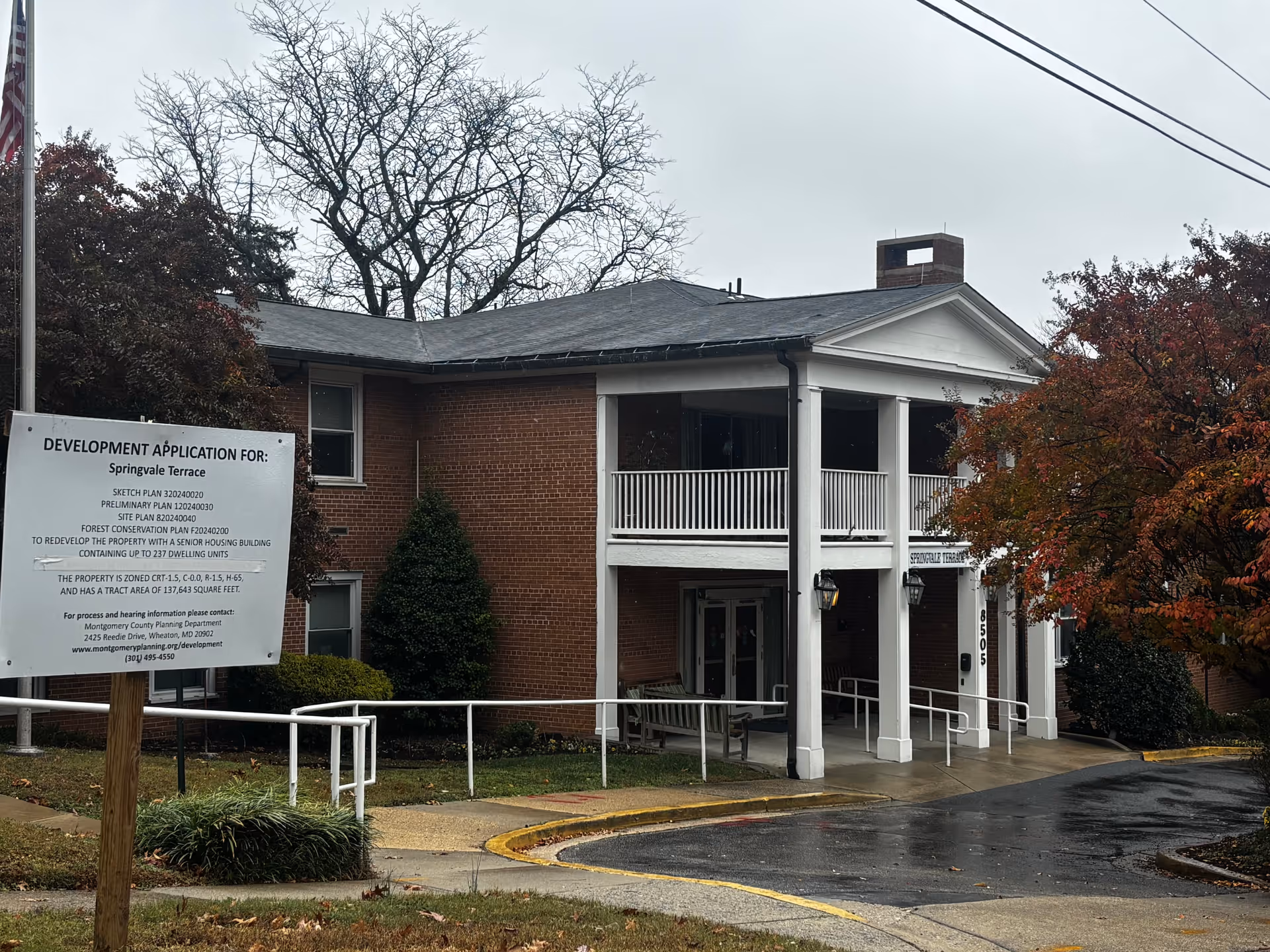 Exterior view of a two-story brick building with white columns and railings at the entrance. There is a sign in front of the building indicating a development application for Springvale Terrace. The surrounding area has trees with autumn foliage and a wet driveway.
