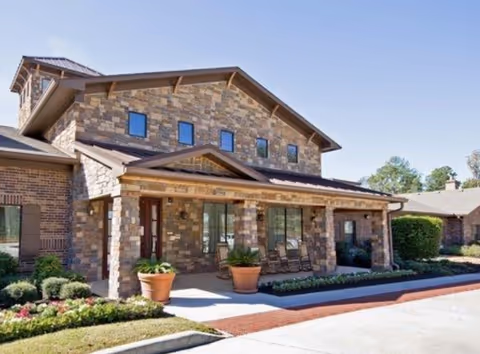 Stone-clad assisted living building front with porch, planters, and landscaped entrance.