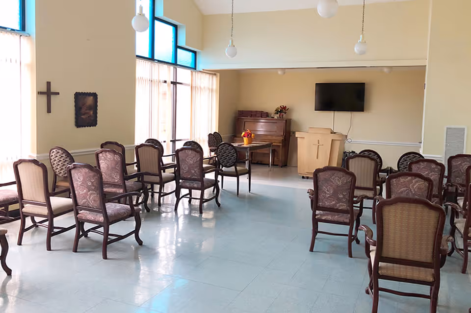 A small chapel or meeting room with rows of upholstered wooden chairs arranged facing a wooden podium with a cross on it. There is a piano and a table with flowers in the corner, a wall-mounted TV above the podium, and large windows with sheer curtains letting in natural light. The walls are light-colored and there are hanging globe light fixtures from the ceiling.