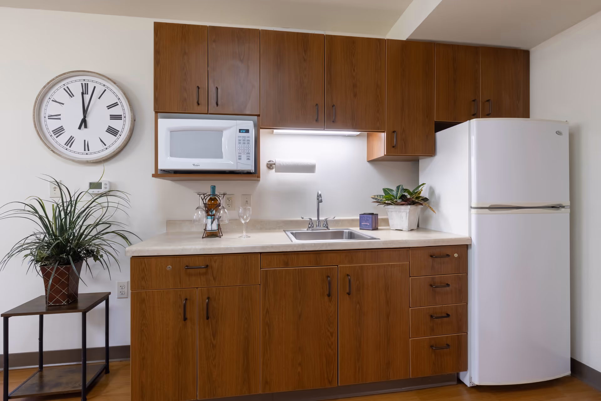 A small kitchen area with wooden cabinets, a white microwave, a white refrigerator, a sink, and a countertop with a wine bottle, two wine glasses, and a potted plant. A large round wall clock and a potted plant on a small table are visible to the left.