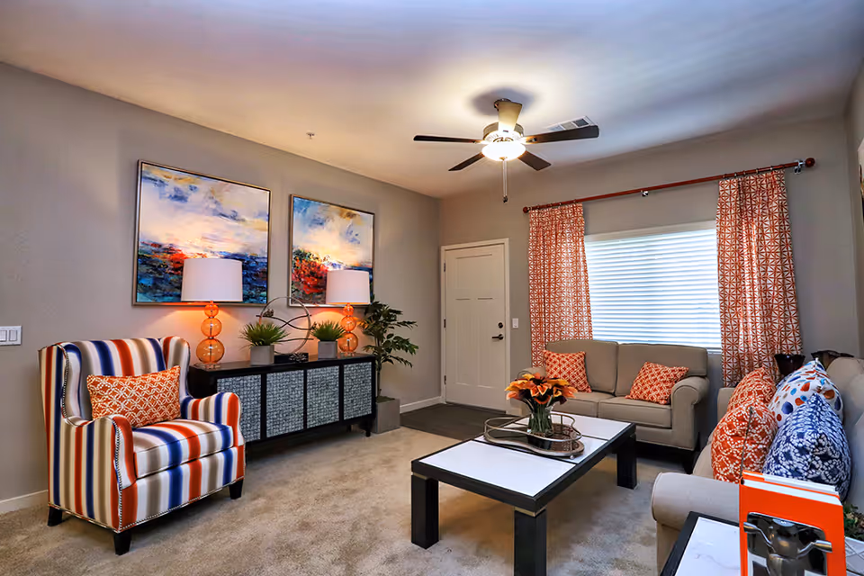 Bright living room with a striped armchair, two sofas, a coffee table, colorful artwork, lamps, and a ceiling fan.