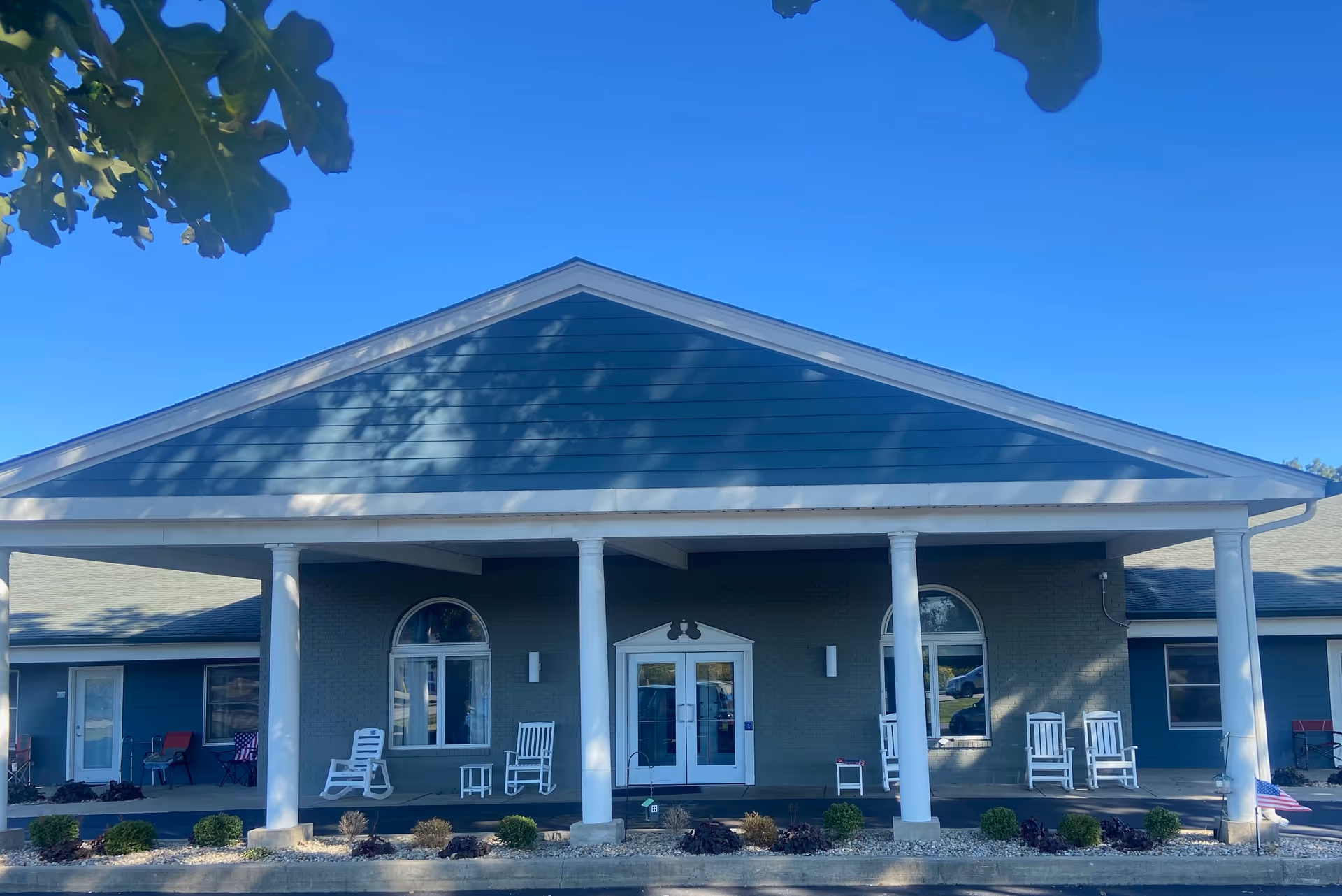 Front exterior view of a single-story building with a large covered porch supported by white columns. The building has blue siding and a gray roof, with several white rocking chairs and small tables arranged on the porch. There are two arched windows and a double glass door entrance. Small shrubs and landscaping are visible in front of the porch under a clear blue sky.