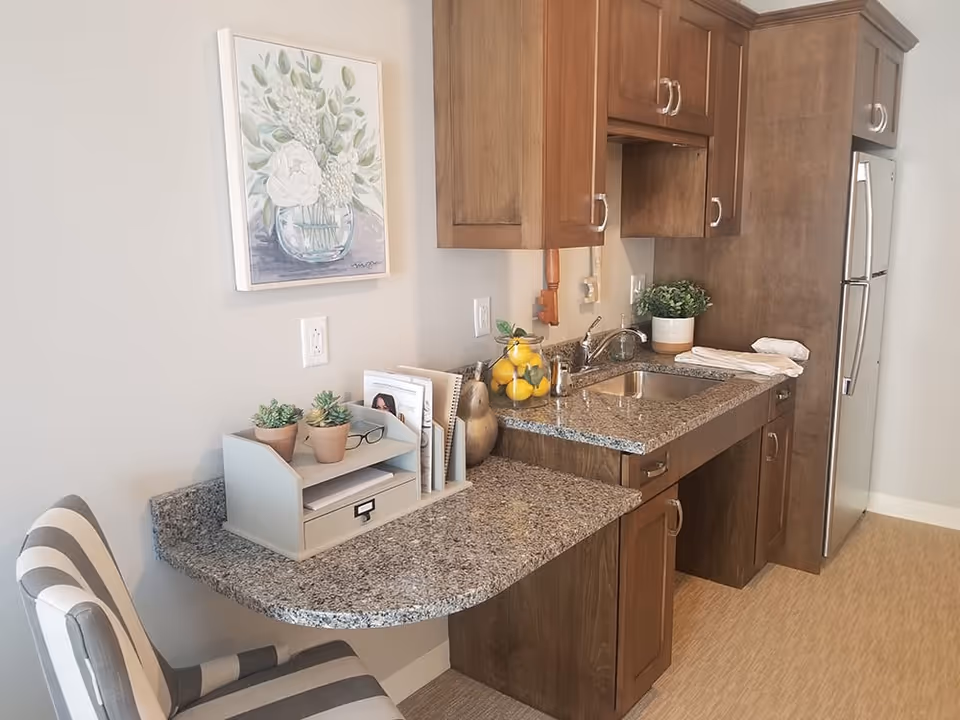 Small kitchenette with granite countertop, wooden cabinets, sink, stainless refrigerator and a striped chair.