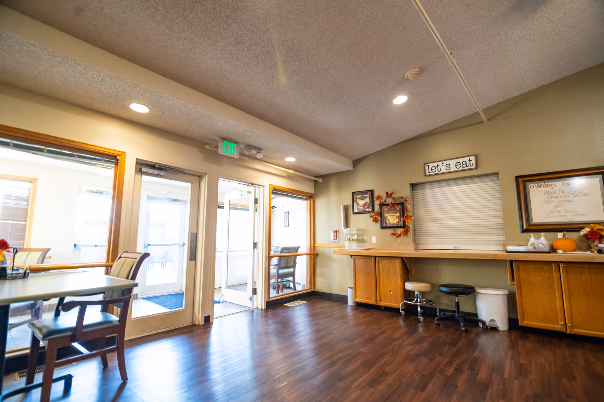 Interior view of a dining area in a senior living facility with wooden floors, tables and chairs, a counter with stools, and a sign on the wall that says 'let's eat'. There are large windows and glass doors allowing natural light to enter the room.