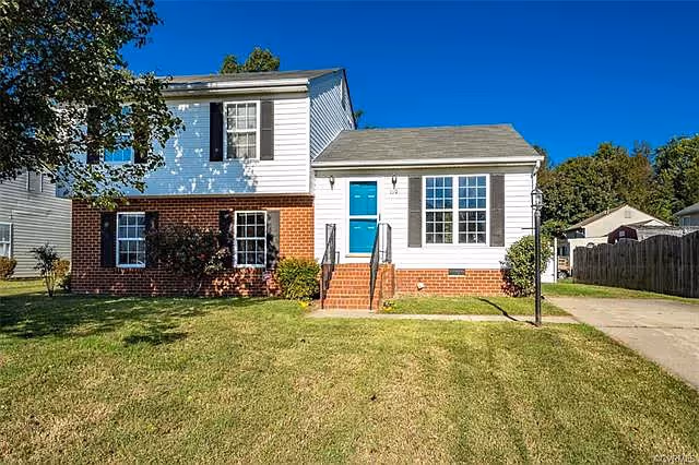 Exterior view of a two-story residential building with white siding and red brick on the lower half. The front door is painted bright blue and is accessed by a small set of brick steps with black railings. There are several windows, a driveway to the right, and a well-maintained lawn in front under a clear blue sky.