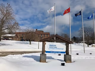 Snow-covered ground with a wooden sign reading 'Atrium Senior Living of the Northland Lodge' and the address '2259 Garfield Street' in front of a brick building. Four flagpoles with flags are visible behind the sign, and leafless trees are scattered around the area under a partly cloudy sky.