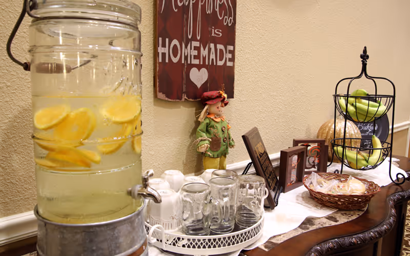 A beverage dispenser filled with lemon water next to a tray holding several clear glasses and white mugs on a wooden table. Nearby, there is a decorative scarecrow doll, a basket with wrapped snacks, a two-tiered fruit basket with green apples and bananas, and a sign on the wall that reads 'Happiness is Homemade' with a heart symbol.