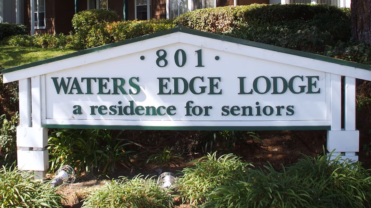 Outdoor sign for Waters Edge Lodge, a residence for seniors, with the address number 801 displayed above the name. The sign is white with green lettering and surrounded by greenery and bushes.