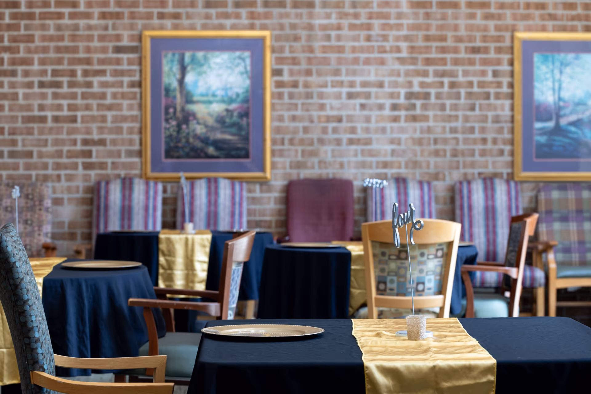 A dining room with several tables covered in black tablecloths and gold runners. Each table has a decorative centerpiece with the word 'hope'. The room has a brick wall with framed landscape paintings and various upholstered chairs arranged around the tables.