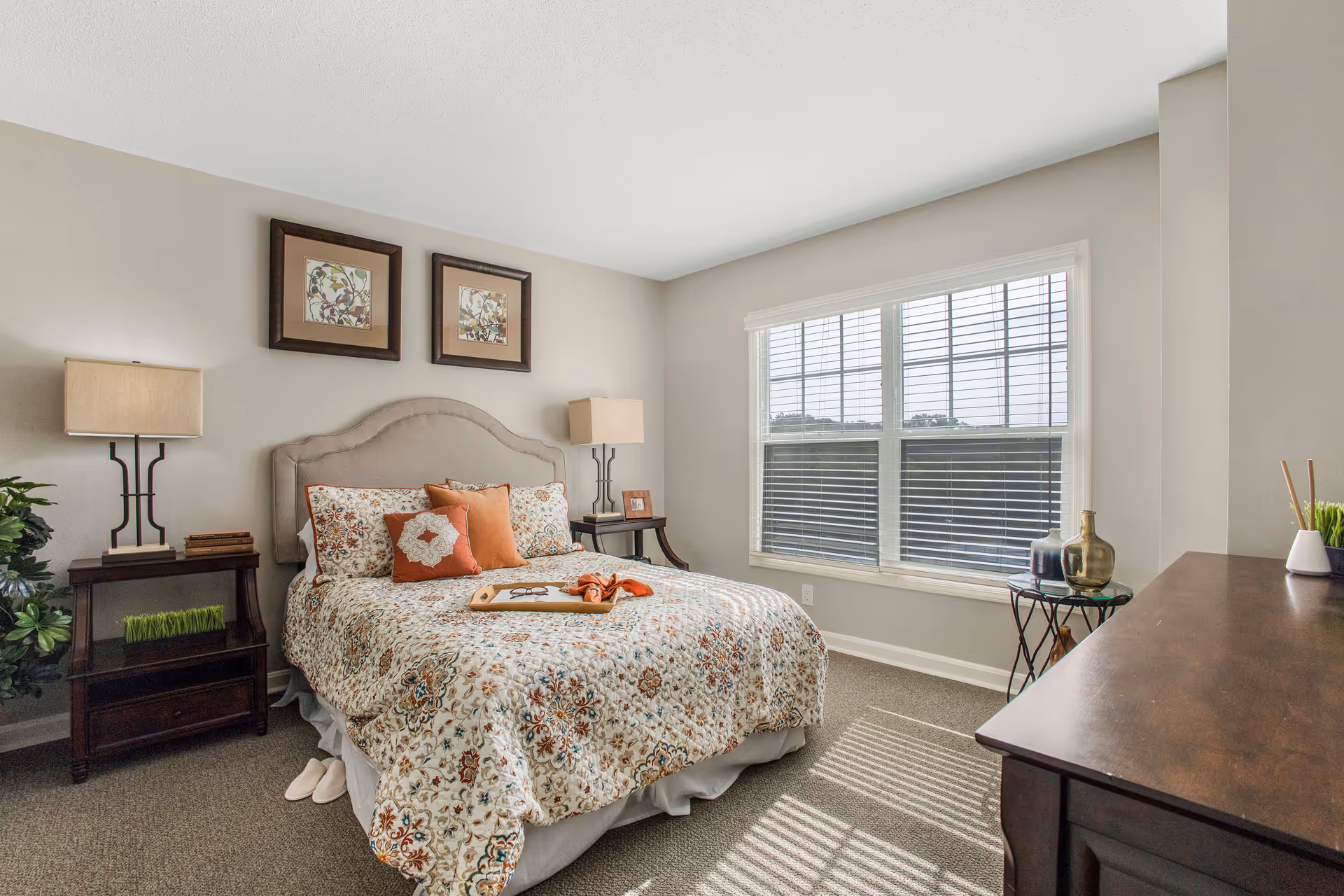 Well-lit bedroom with a patterned bed, two bedside tables with lamps, wall art above the headboard, and a large window with blinds.