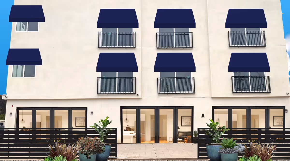 Exterior view of a modern multi-story building with beige walls and multiple windows, each topped with dark blue awnings. The ground floor features large glass doors leading to interior rooms, with a black fence and several potted plants in front. The sky is clear and blue.