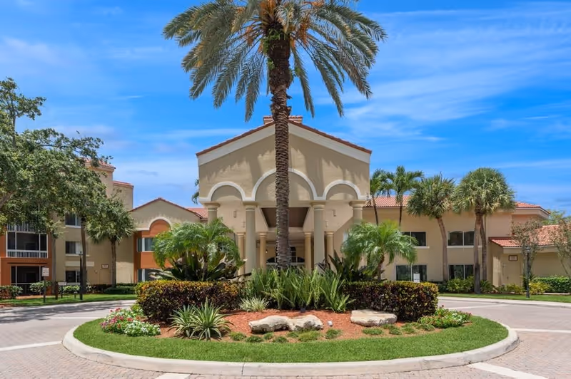 Front exterior view of a senior living facility with a circular driveway, landscaped garden with palm trees and shrubs, and a beige building with red-tiled roof under a blue sky.