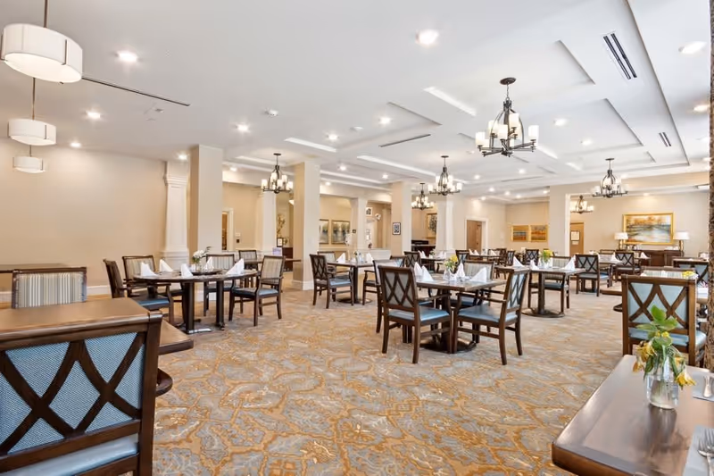Spacious dining room with multiple wooden tables and chairs arranged neatly. Each table is set with white napkins and small flower vases. The room features beige walls, patterned carpet, and several chandeliers hanging from a white ceiling with recessed lighting.