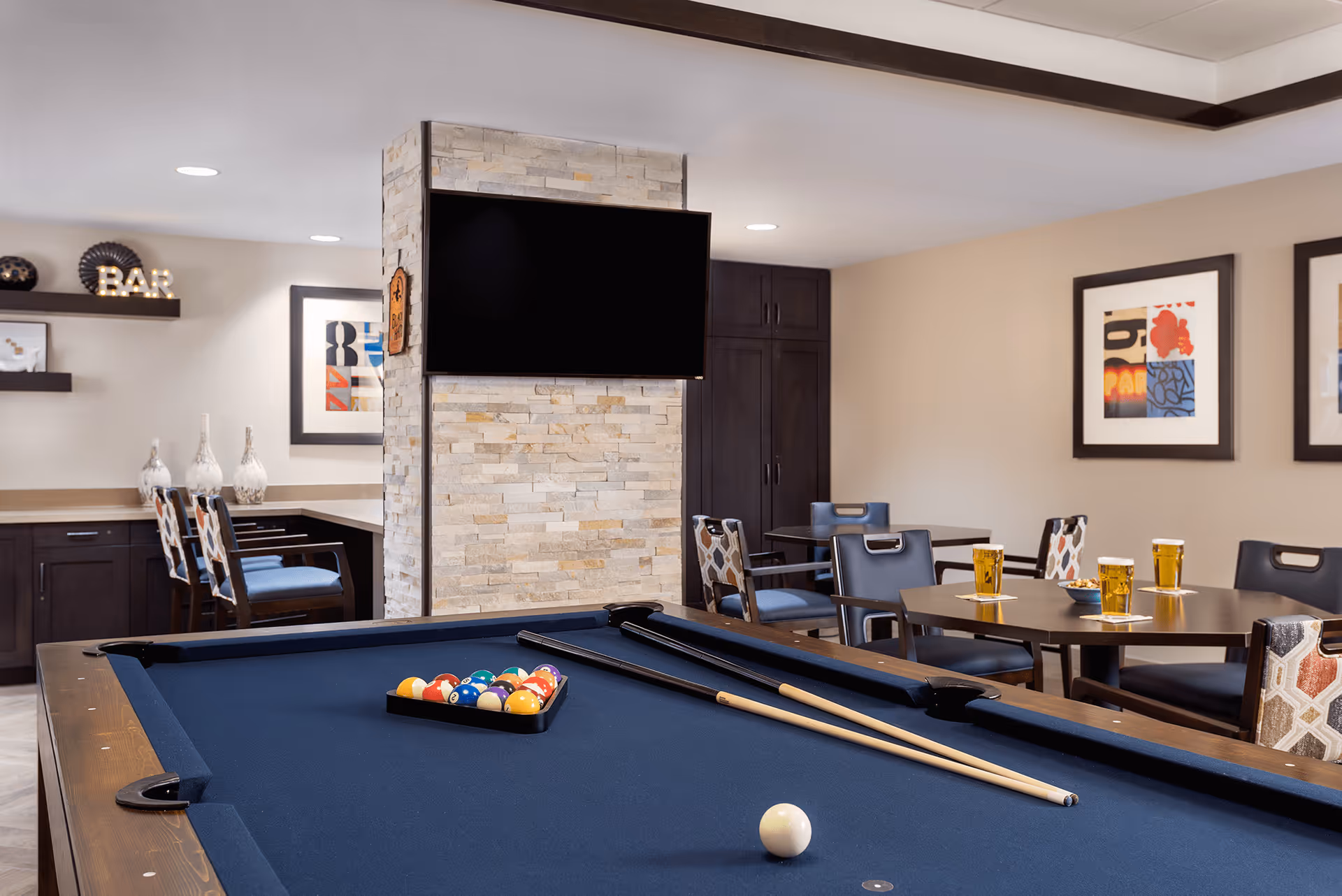A recreational room with a pool table in the foreground, two pool cues resting on the table, and a rack of billiard balls. In the background, there are tables with chairs, some with glasses of beer, a wall-mounted flat-screen TV above a stone pillar, and decorative framed artwork on the walls. Shelves and cabinets are visible along the back wall.