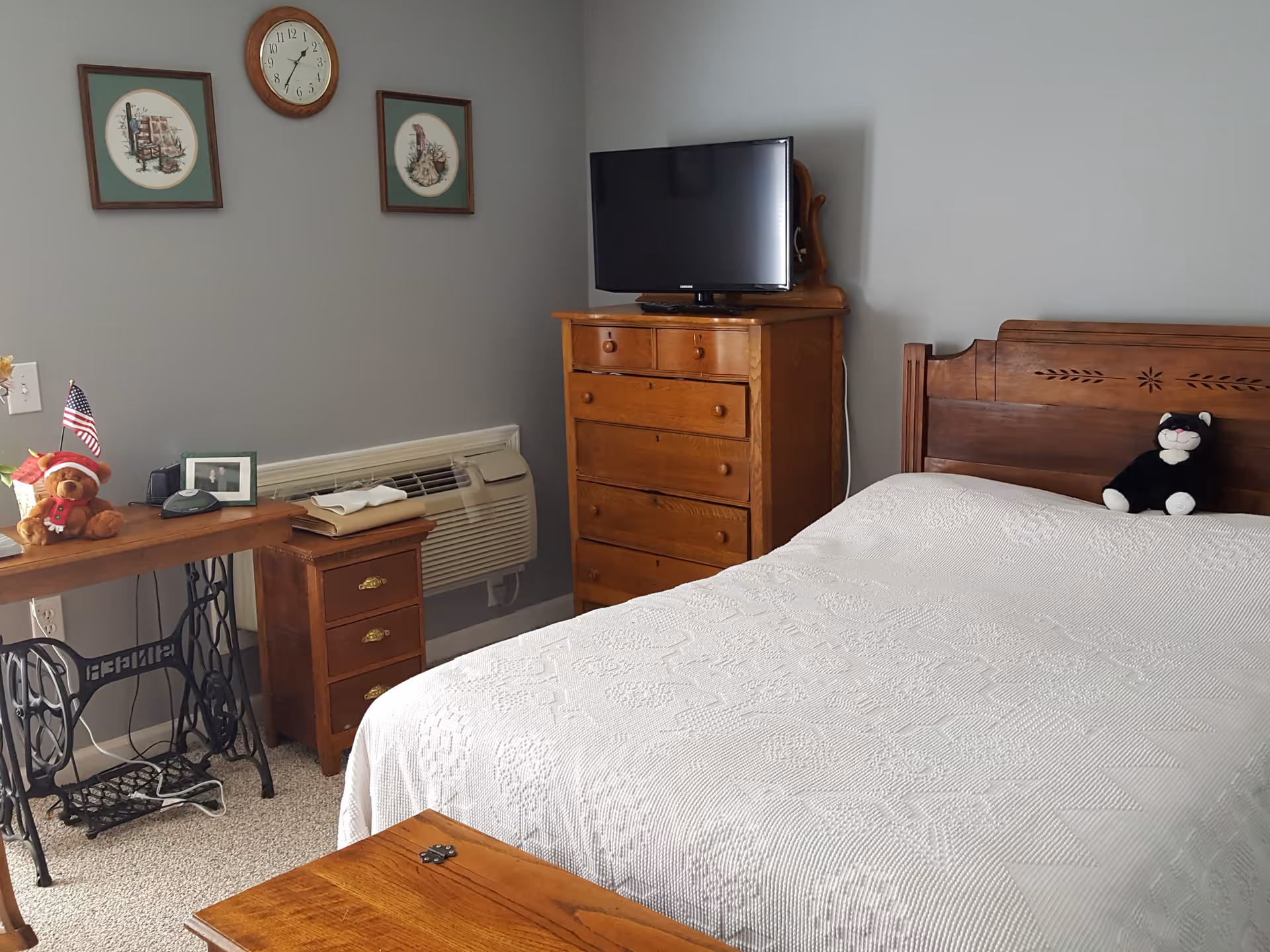A bedroom with a wooden bed frame and a white textured bedspread. A black and white stuffed cat sits on the bed. There is a wooden dresser with a flat-screen TV on top. On the left side, there is a small wooden table with a teddy bear holding an American flag, a framed photo, and a small device. Above the table, a wall clock and two framed pictures hang on a gray wall. An air conditioning unit is installed below the pictures.