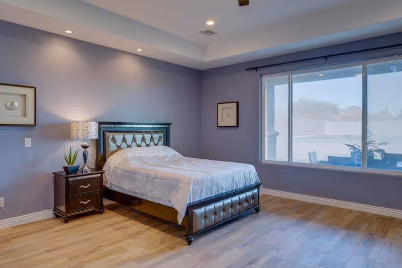 A bedroom with a single bed featuring a tufted headboard and footboard, a nightstand with a lamp and a small plant, light purple walls, wooden flooring, and a large window showing an outdoor patio area.