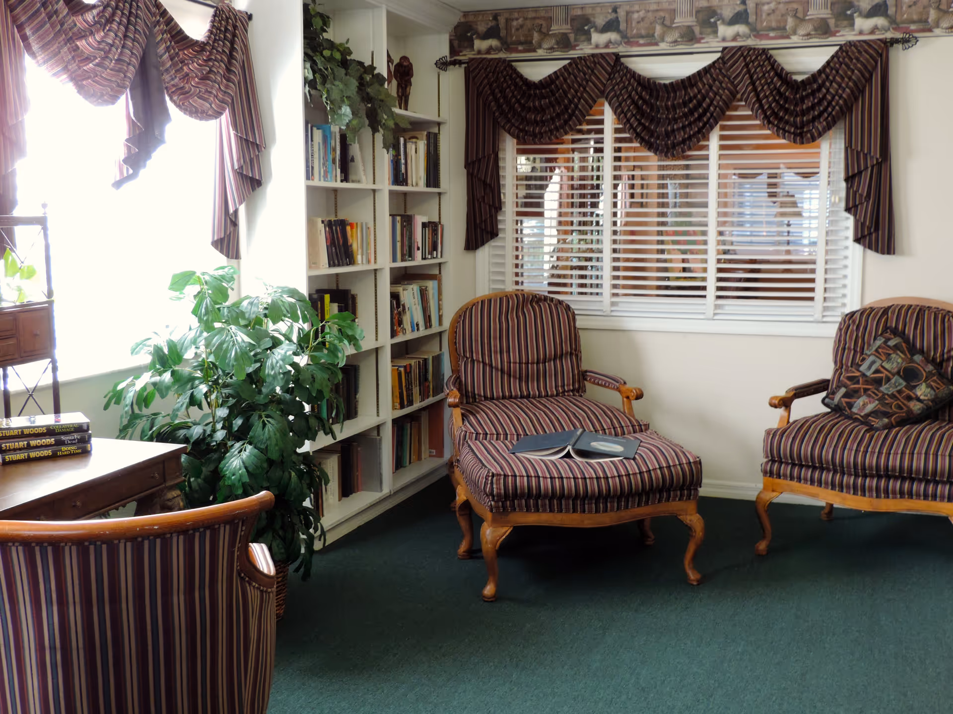 A cozy sitting area with two striped armchairs and a matching ottoman, a wooden desk with books on it, a large leafy plant, a white bookshelf filled with books, and windows with striped valances and white blinds.