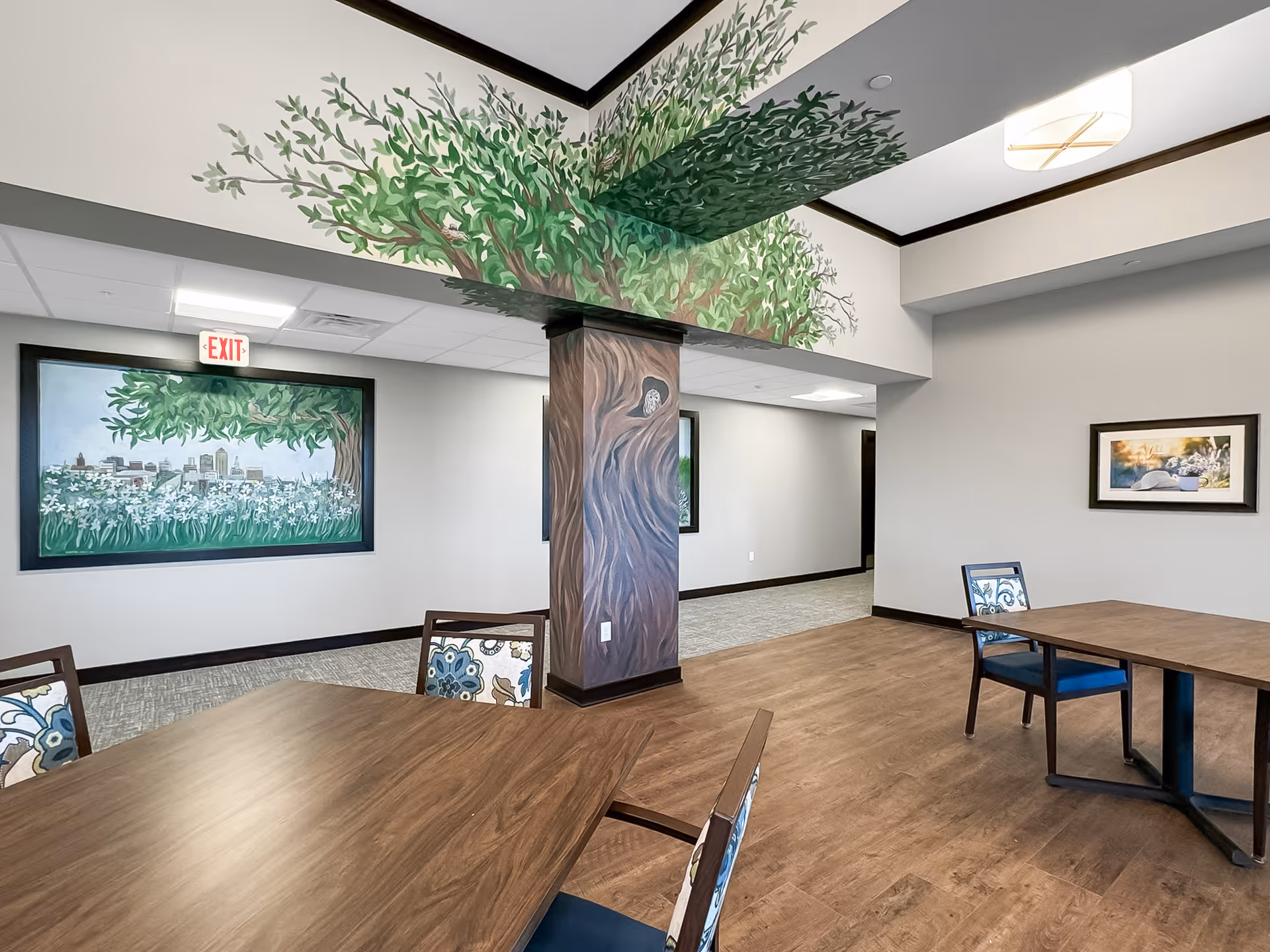 Interior view of a senior living facility common area with wooden tables and chairs featuring floral upholstery. The walls are decorated with murals of trees and flowers, including a tree painted on a column extending onto the ceiling. There are framed pictures on the walls and an exit sign above a doorway.