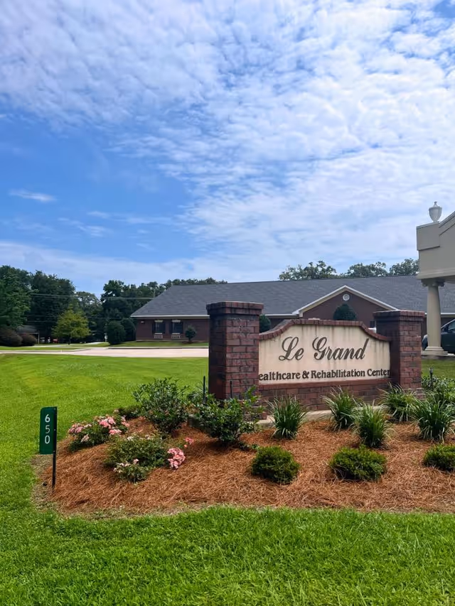 Brick entrance sign reading "Le Grand Healthcare & Rehabilitation Center" in a landscaped lawn with the facility building and blue sky in the background.