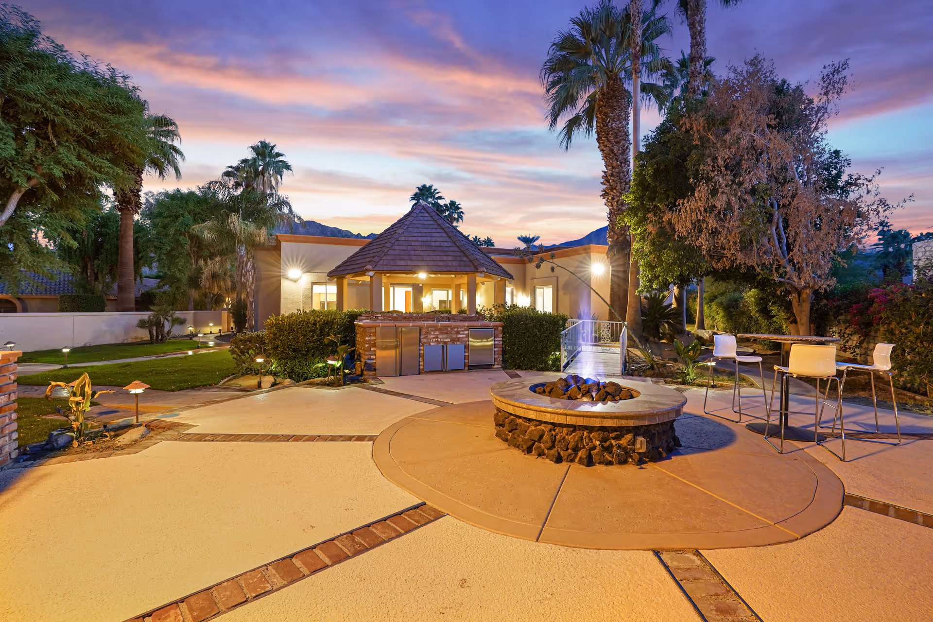 Outdoor patio area at sunset with a round stone fire pit in the center, surrounded by a paved surface with brick accents. There is a small gazebo-like structure with a shingled roof and built-in outdoor kitchen appliances. Several palm trees and other greenery surround the area, with a table and chairs set up for seating.