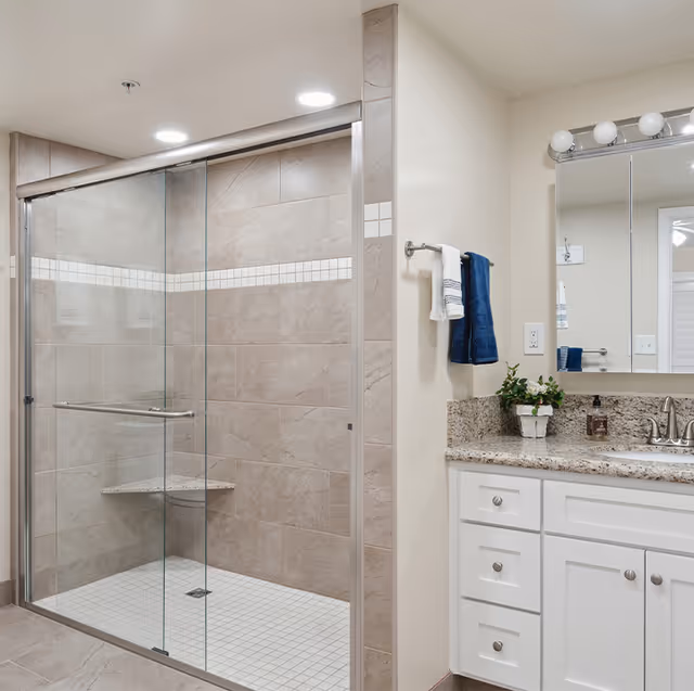 A modern bathroom featuring a large glass-enclosed shower with beige tiles and a built-in corner seat. Next to the shower is a white vanity with granite countertop, a sink, a mirror with lights above, and a towel rack holding a white and a blue towel. A small potted plant and soap dispenser are on the countertop.