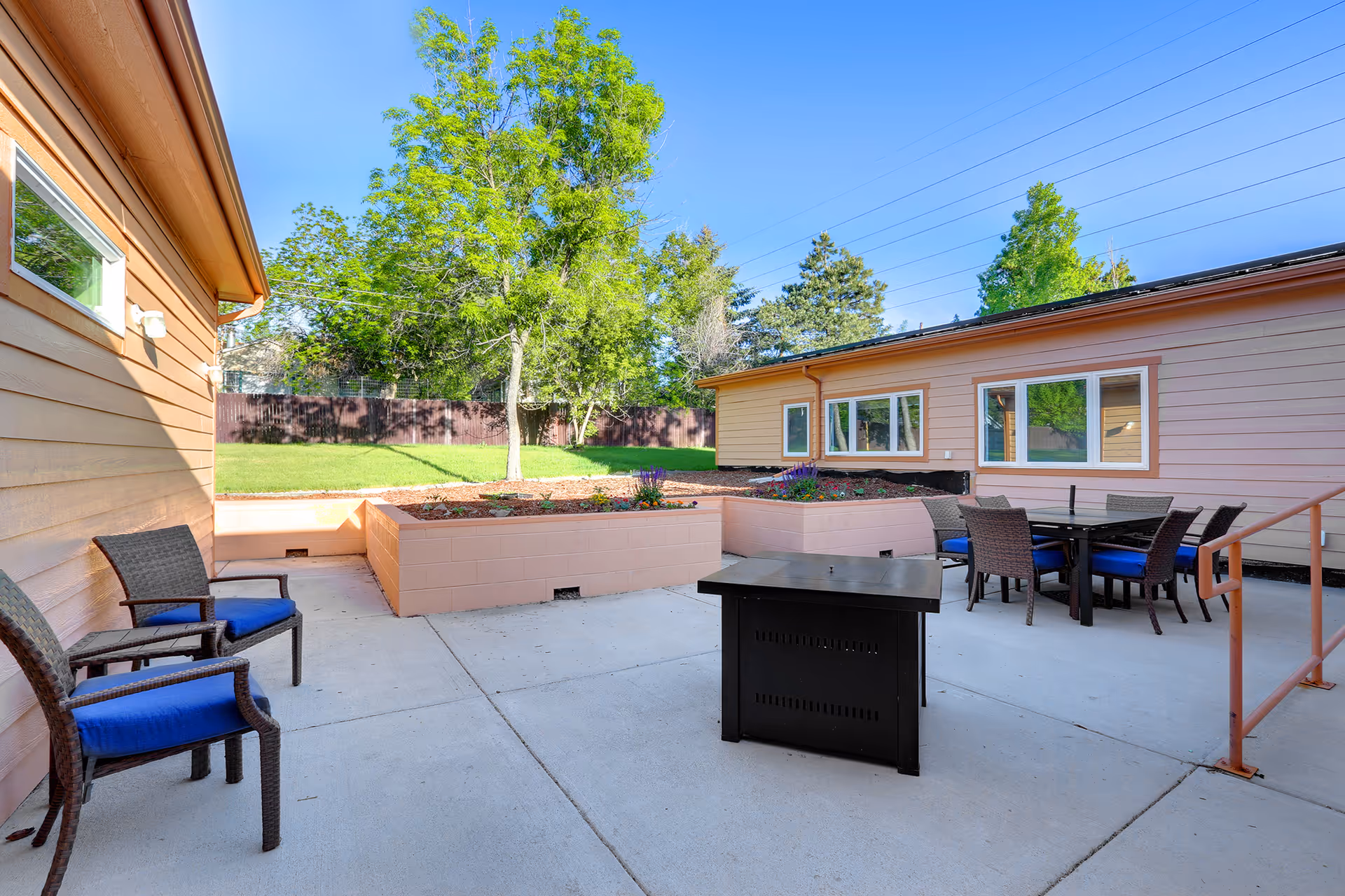 Outdoor patio area with several wicker chairs with blue cushions, a black fire pit table, and a dining table with chairs. Raised garden beds with flowers and a grassy area with trees and a wooden fence are visible in the background under a clear blue sky.