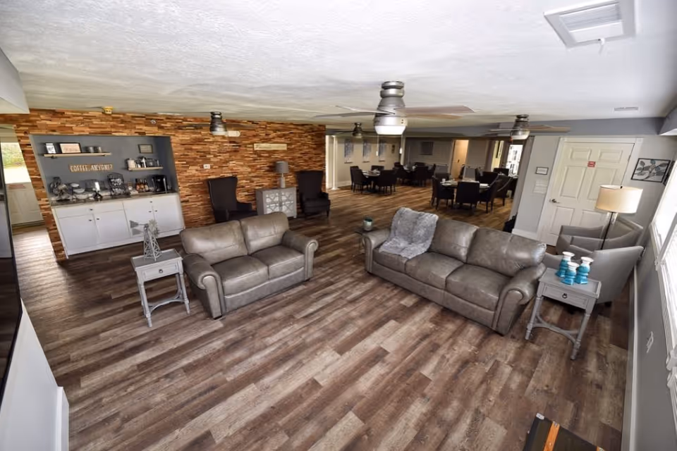 A spacious senior living common area with two gray leather sofas, two gray armchairs, and side tables. The room features wood flooring, ceiling fans, and a coffee station against a wooden accent wall. In the background, there are several dining tables and chairs arranged in a dining area.