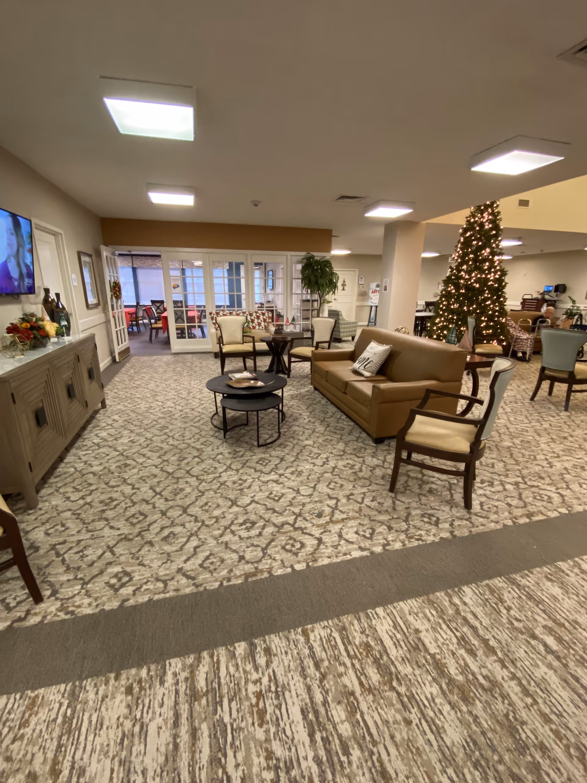A spacious senior living facility common area with patterned carpet flooring, a brown leather sofa, several cushioned chairs, and two round black nesting coffee tables. A decorated Christmas tree with lights stands near the center. In the background, there is a glass door leading to another room with red chairs and tables. The room is well-lit with ceiling lights, and a TV is mounted on the left wall above a wooden cabinet.