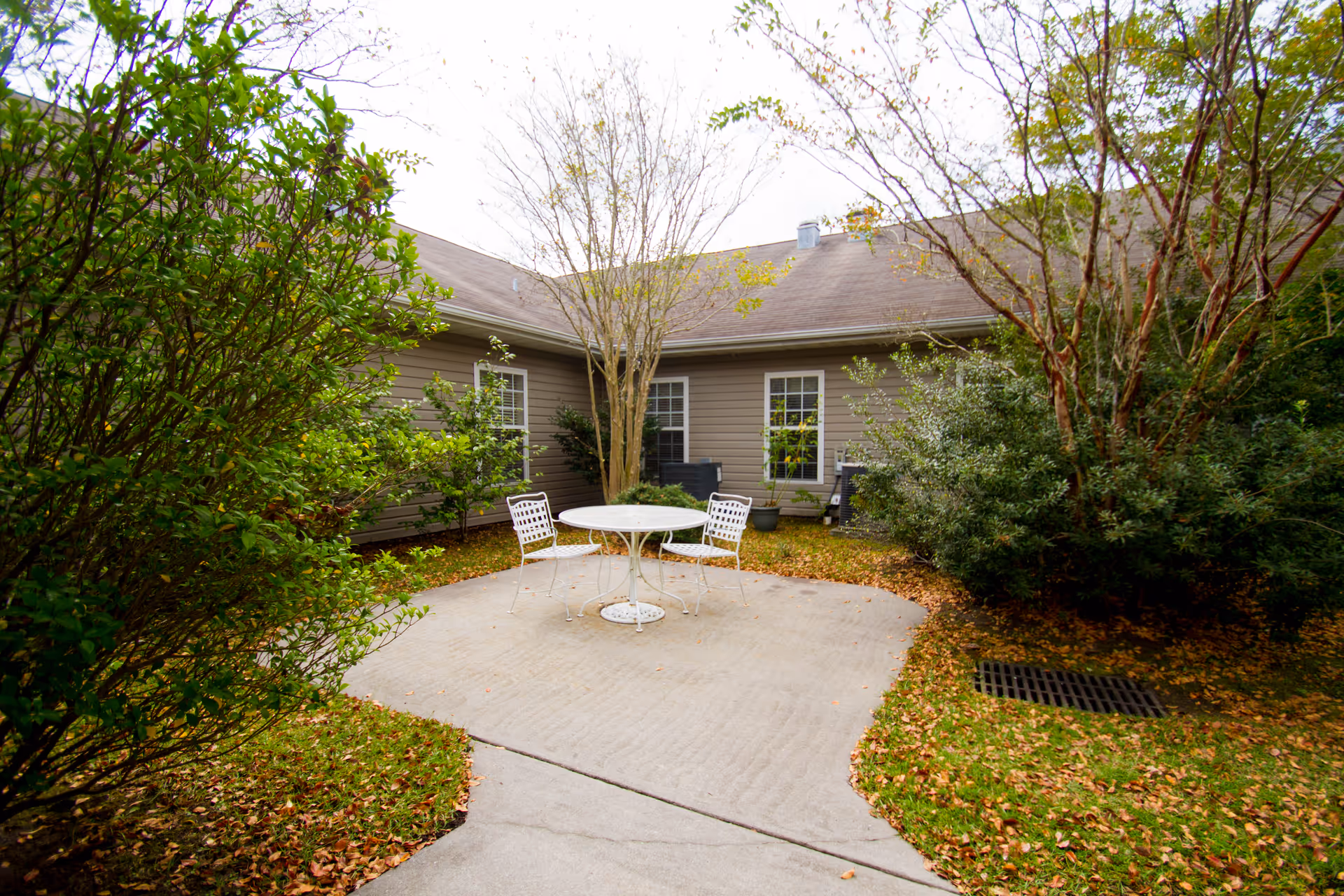 Outdoor patio area with a round white metal table and two matching chairs on a concrete slab surrounded by green bushes and trees with a building in the background.