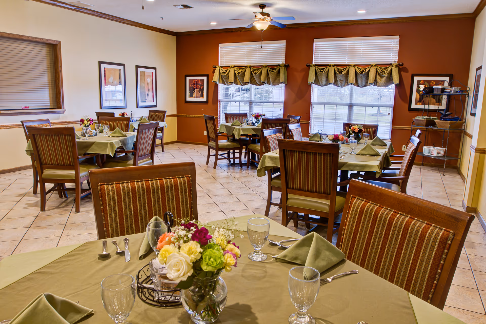 A dining room with several tables covered in green tablecloths and set with glassware, silverware, and floral centerpieces. The room has tiled floors, large windows with green valances, framed artwork on the walls, and a ceiling fan with a light fixture.