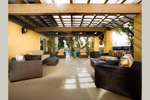 Covered outdoor patio with wicker sofas and striped cushions under a wooden pergola and potted plants.