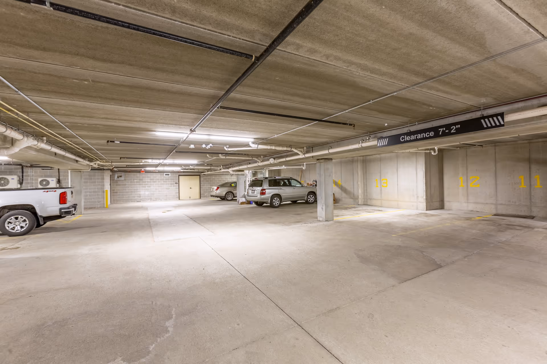 Indoor parking garage with concrete floors, walls, and ceiling. Several cars are parked in designated spaces marked with yellow numbers. A clearance sign indicating 7 feet 2 inches hangs from the ceiling.